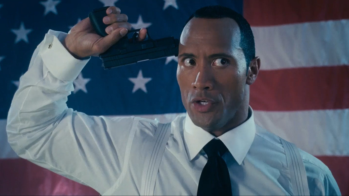 Dwayne Johnson holds a gun to his head in front of the American flag in Southland Tales.