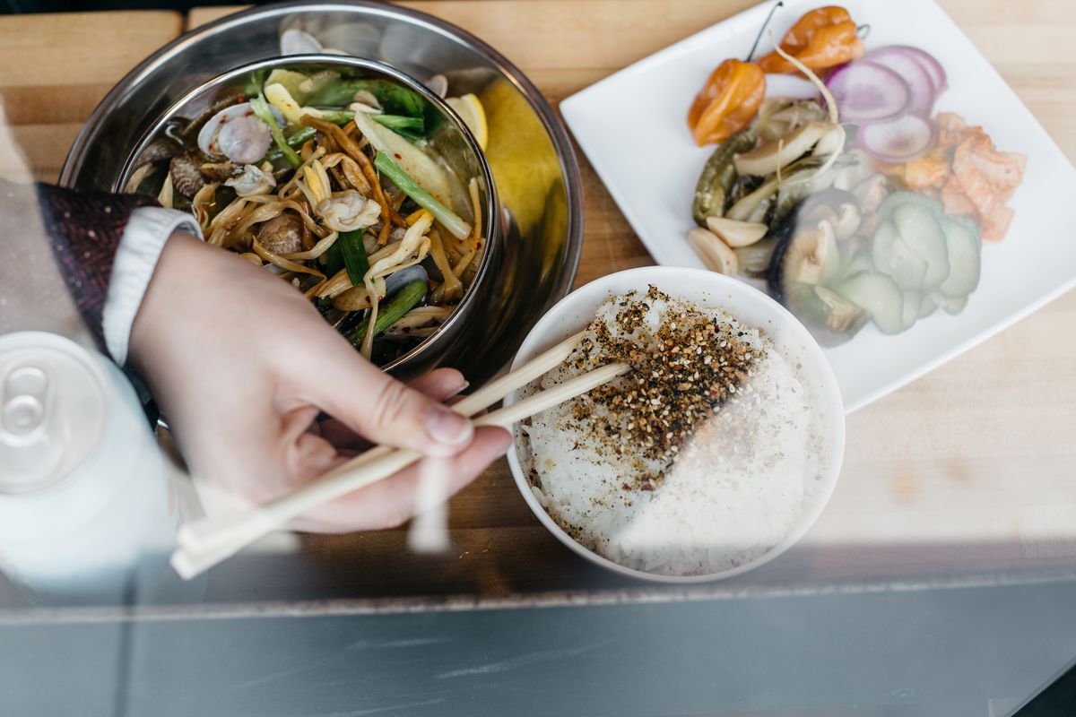 A hand with chopsticks reaches for furikake rice next to a metal bowl of clams and a plate of pickled veggies. 