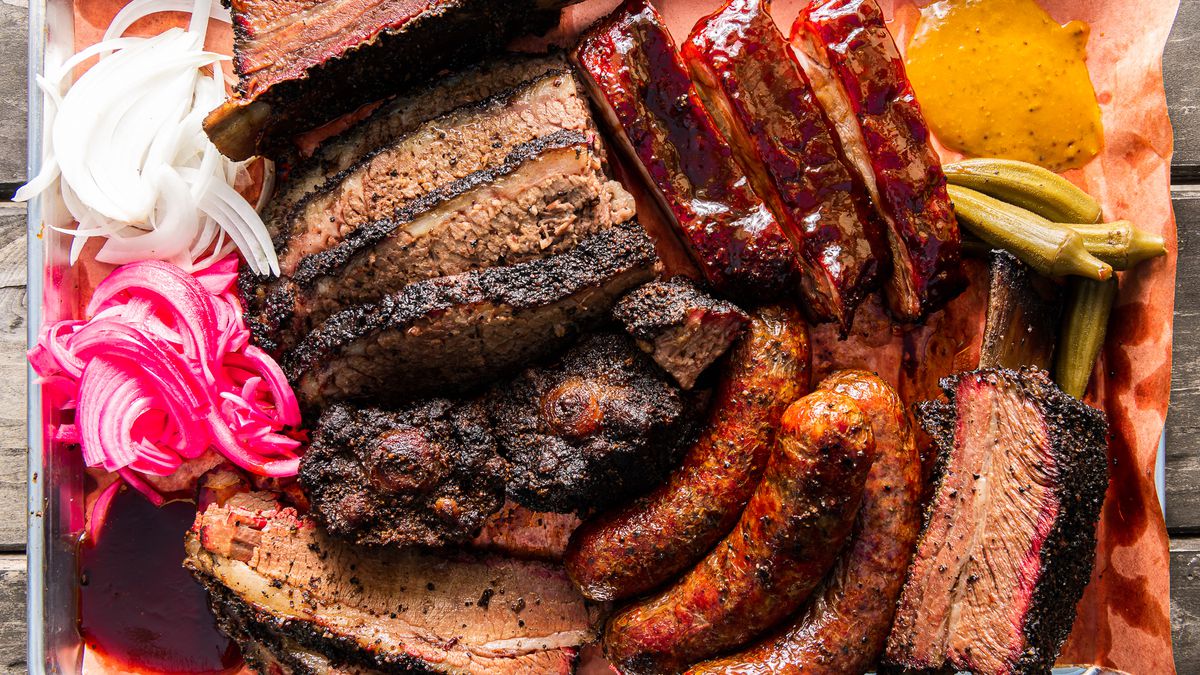 A tray of barbecued meats — sliced brisket, ribs, links, and an on-the-bone beef rib — with a small pile of pink pickled onions to the side