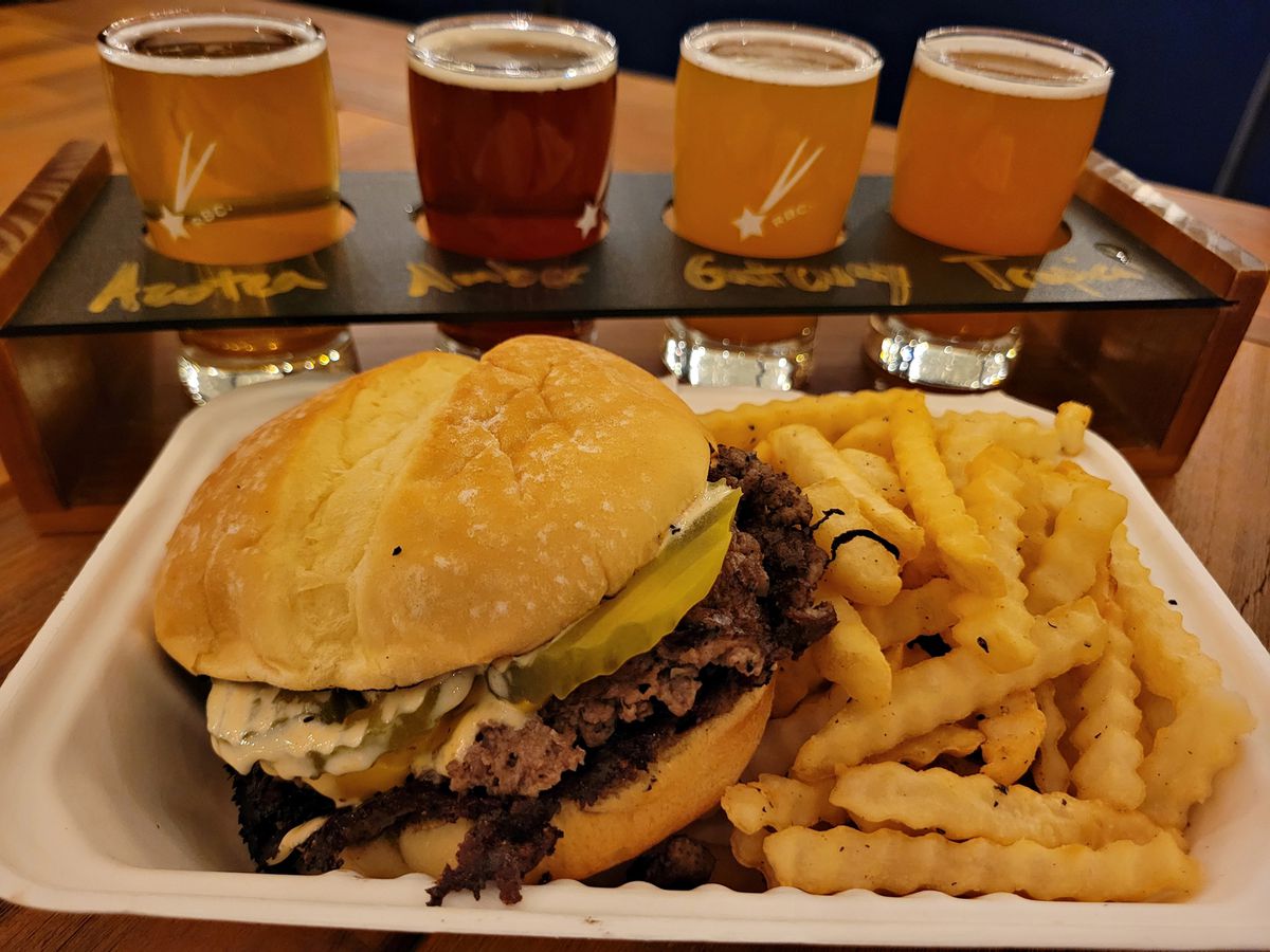 A burger and fries in a styrofoam container next to a flight of beers.