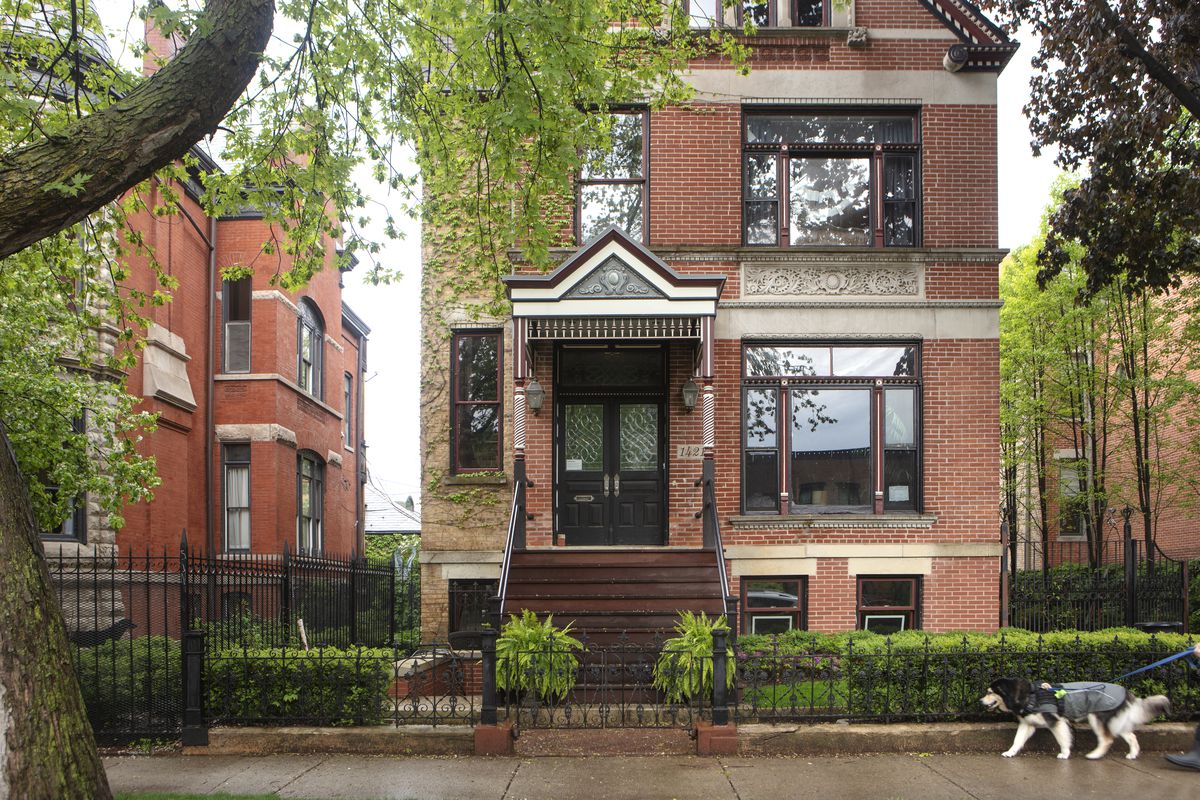 A photo of a two-story brick home. There are manicured bushes, a tall tree, and a woman walking a dog on a sidewalk. It is rainy.