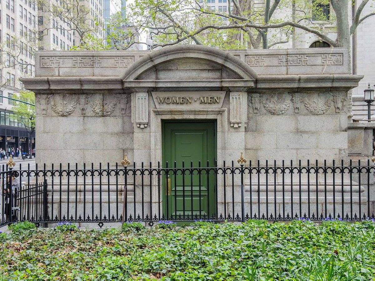 The exterior of a public bathroom in Bryant Park. The facade is concrete and the door is arched.
