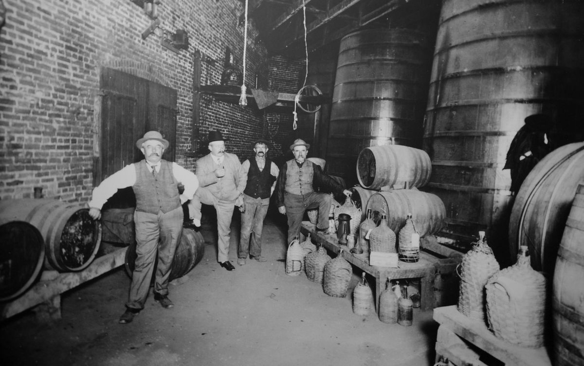A black-and-white vintage photograph shows four men in vests and hats posting near wine barrels and jugs in a windowless room. 