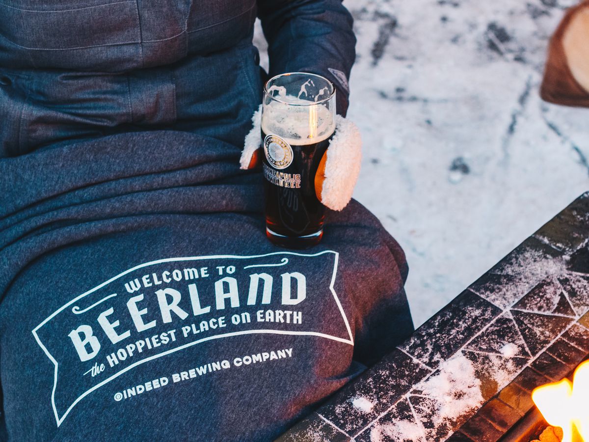 A person sits with a “Beerland: the hoppiest place on earth” t-shirt over their lap. A beer in a mittened hand, and a firepit is visible at the edge of the image.