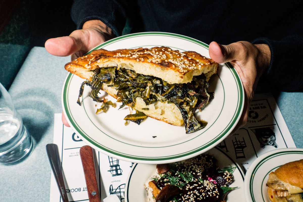 A hand holds a sandwich made from a slice of focaccia overflowing with collard greens.