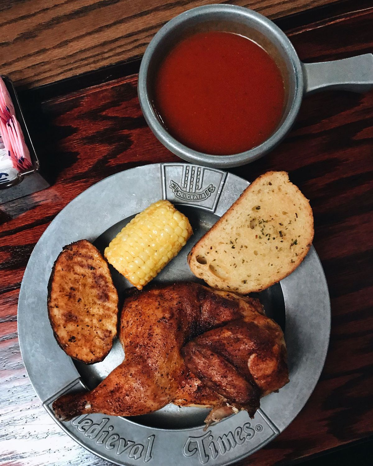 Plate with “Medieval Times” written on it containing a piece of bread, roasted chicken, corn, and half of a potato. A bowl sits alongside containing tomato soup.
