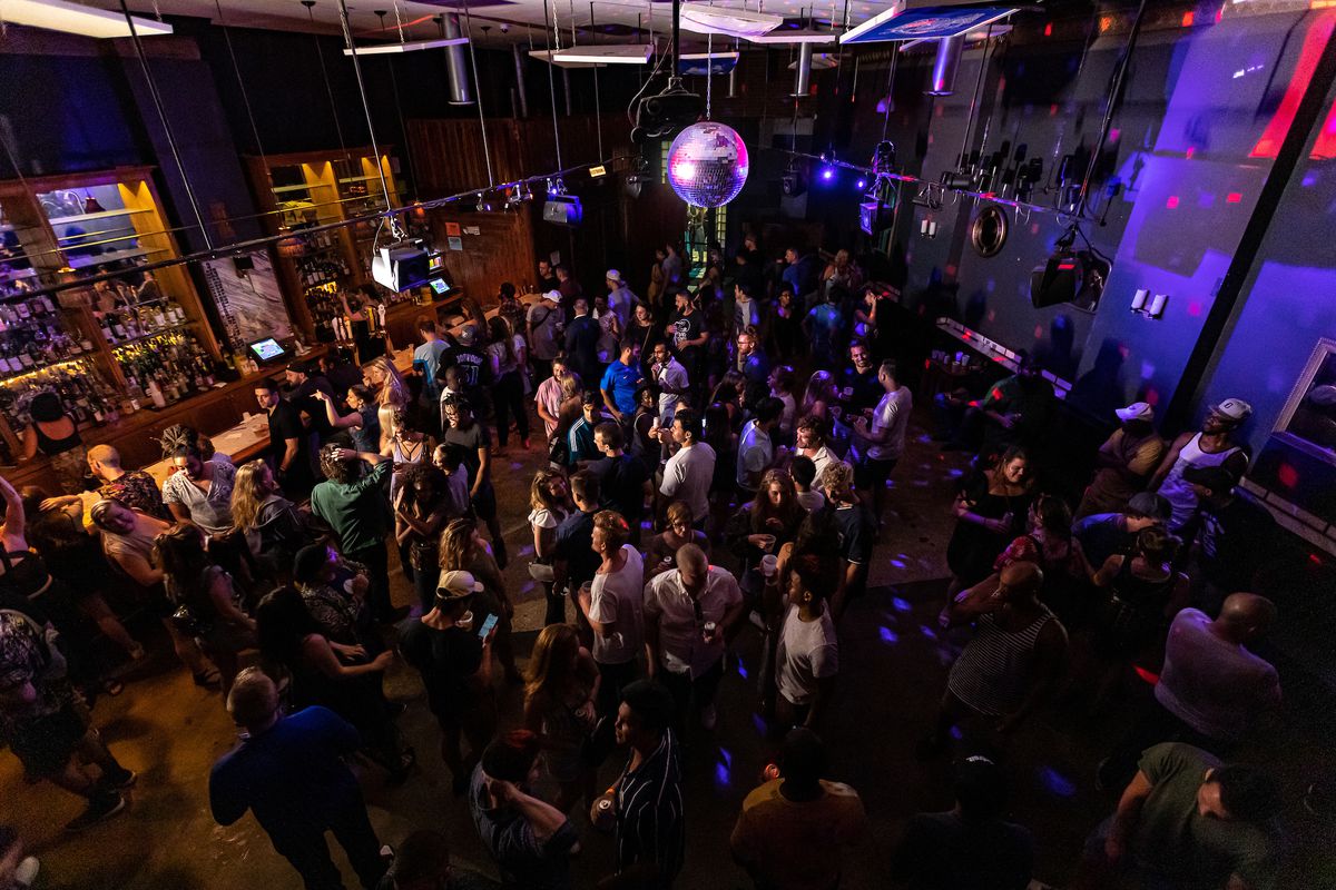 A large group of people gather in a club on the dancefloor beneath a disco ball. 