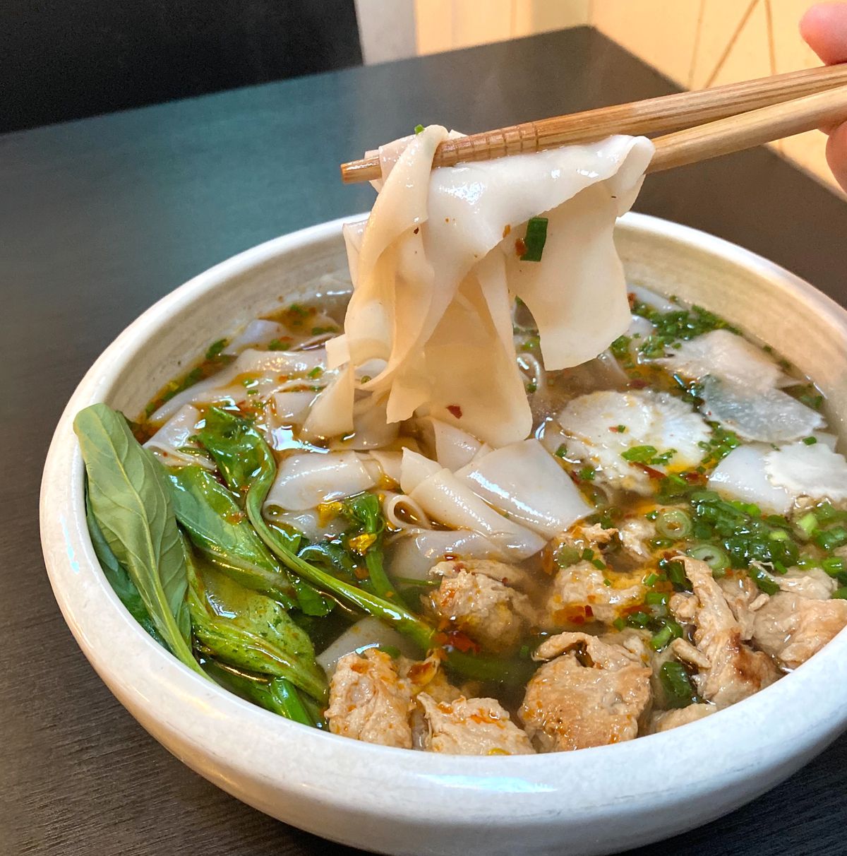 A picture of chopsticks holding up wide rice noodles in a broth with greens and fake chicken