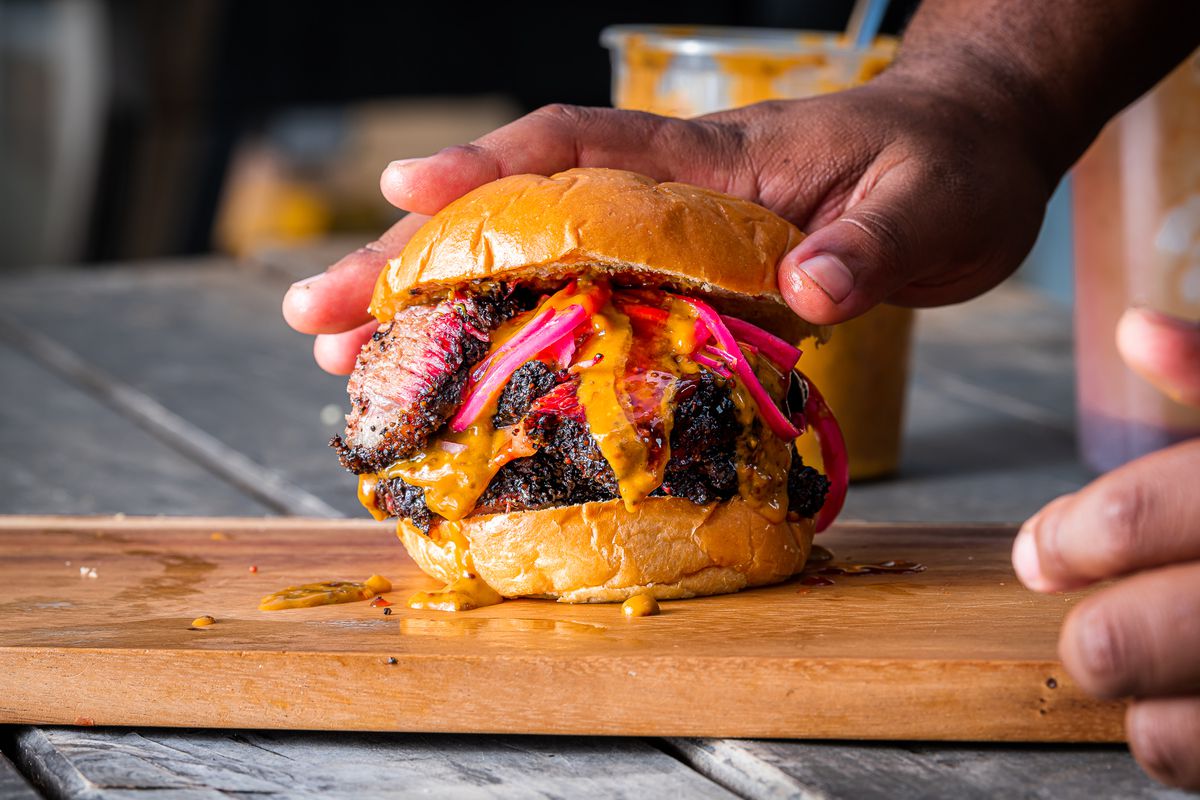 A hand rests on top of a brisket sandwich, the meat piled high on a round bun, with pink pickled onions and golden honey mustard on top