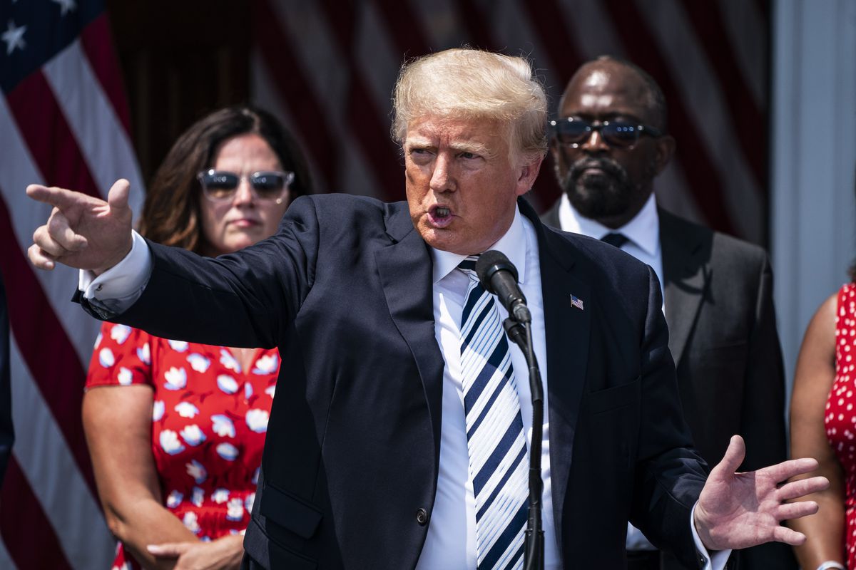 Former President Donald Trump, in a black suit, white shirt, and black-and-silver striped tie, points with one hand and spreads the other open while talking to a crowd outdoors, with supporters and a Secret Service agent standing behind him.