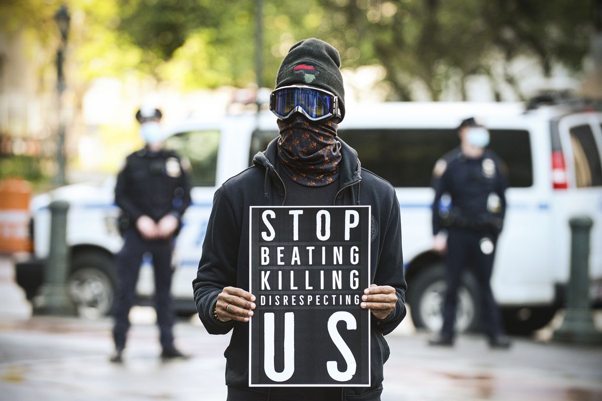 A protester dressed all in black, with a scarf around their face and ski goggles over their eyes, holds a black sign with white letters that read: “Stop beating killing disrespecting us.”