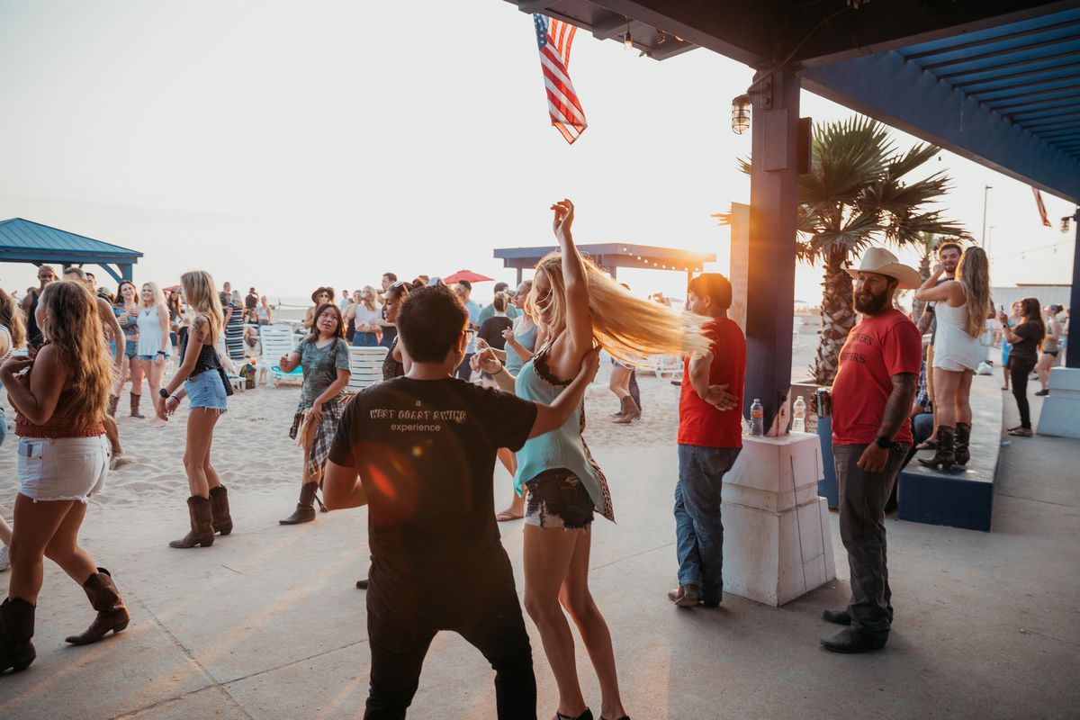Dancers twirl at sunset wearing country and western gear at a beach bar.
