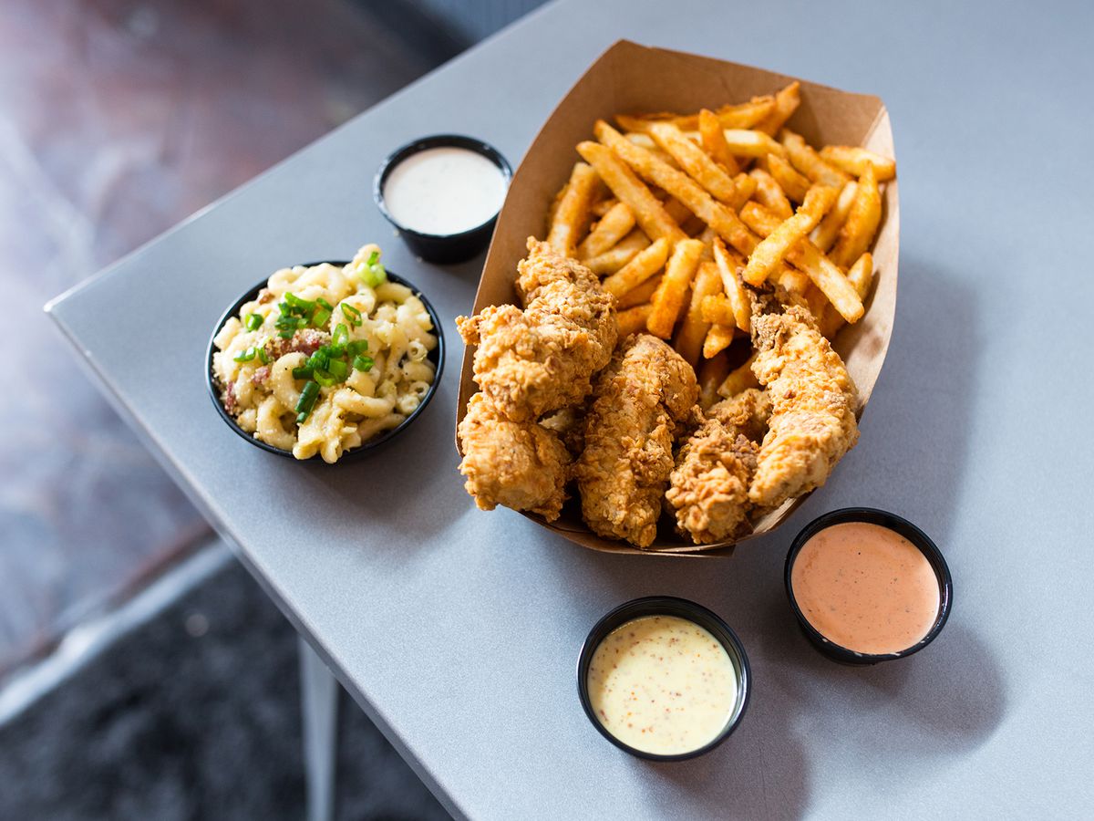 A cardboard paper basket of fries and chicken tenders next to a small bowl of mac and cheese and three saucers of sauces on a table.