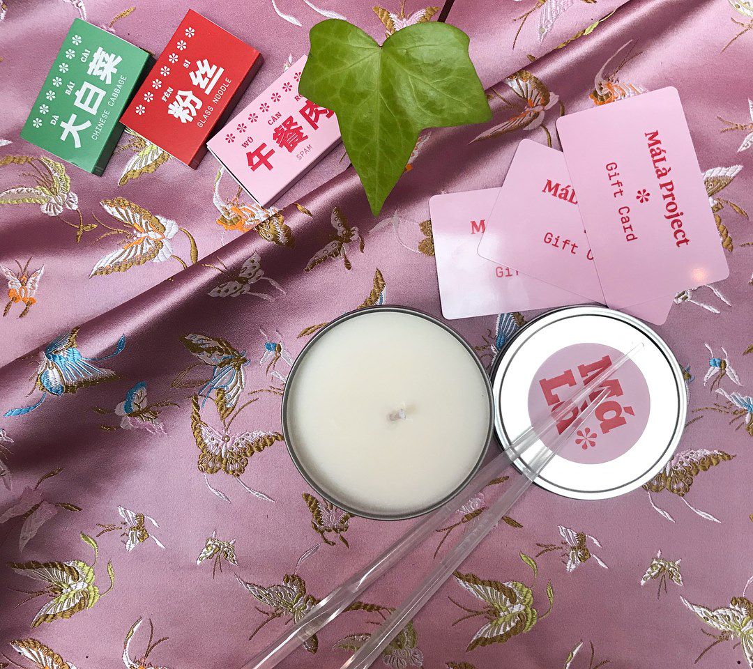 On top of a shiny pink cloth with golden and blue butterfly patterns are a clear pair of acrylic chopsticks that lie on top of the lid of an open candle, and MaLa project gift cards next to a green leaf