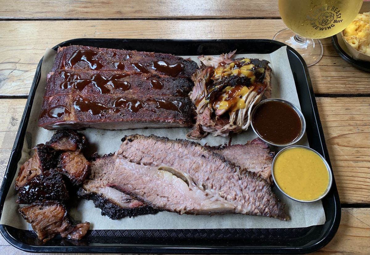A plastic tray with butcher paper holds brisket burnt ends, brisket, two barbecue sauces, pulled pork with sauce, and spare ribs. 