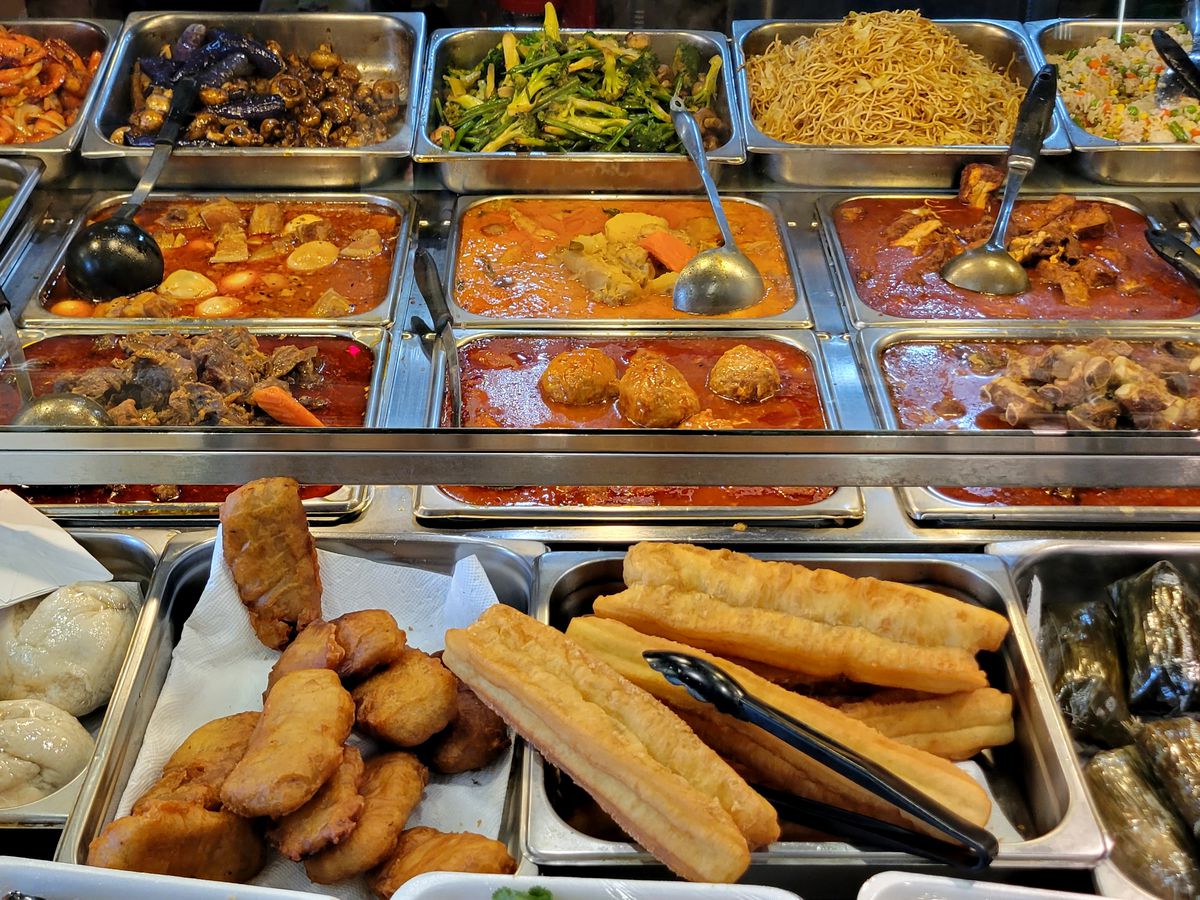 A spread of various soups, vegetable stir-fries, noodle dishes, and other foods in metal trays or containers in a deli.