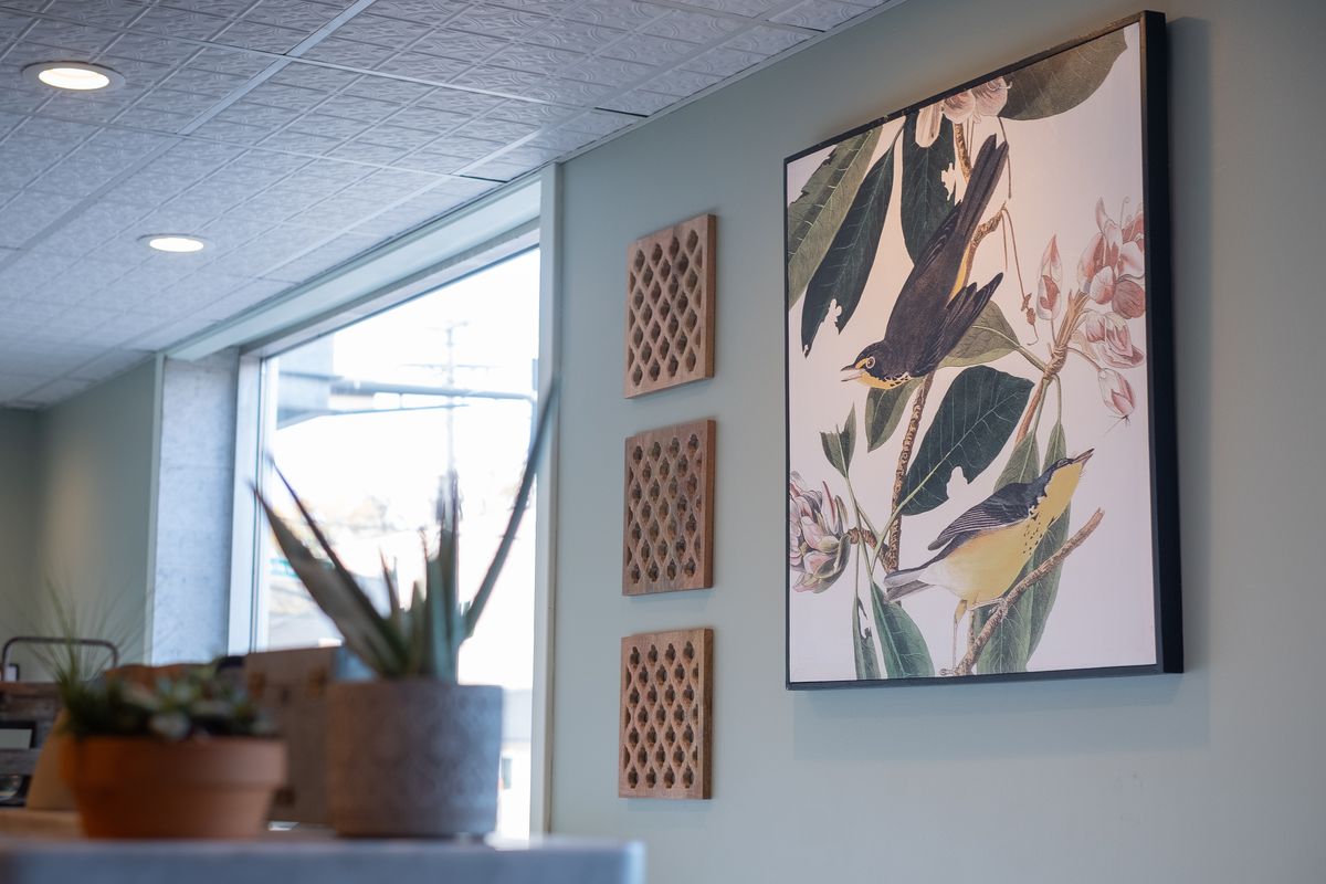 A pale green wall is decorated with a bird painting. Out of focus, there are small green plants near a large window that overlooks St. Clair
