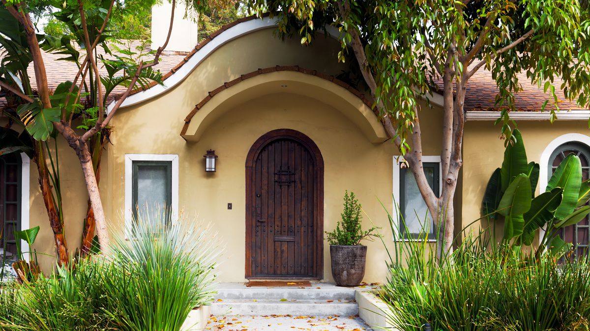 The front door of a home in LA, a rounded wooden doorway, tile roof, surrounded by plants and lush plants. 