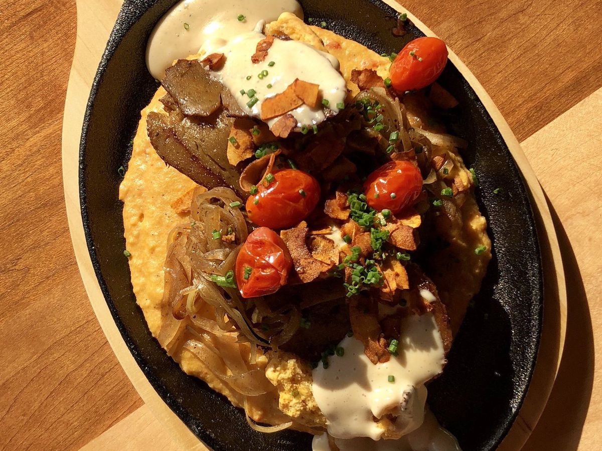 From above, a skillet full of saucy/cheesy looking grits topped with a fried item, crumbled vegan bacon, tomatoes, and other fixings