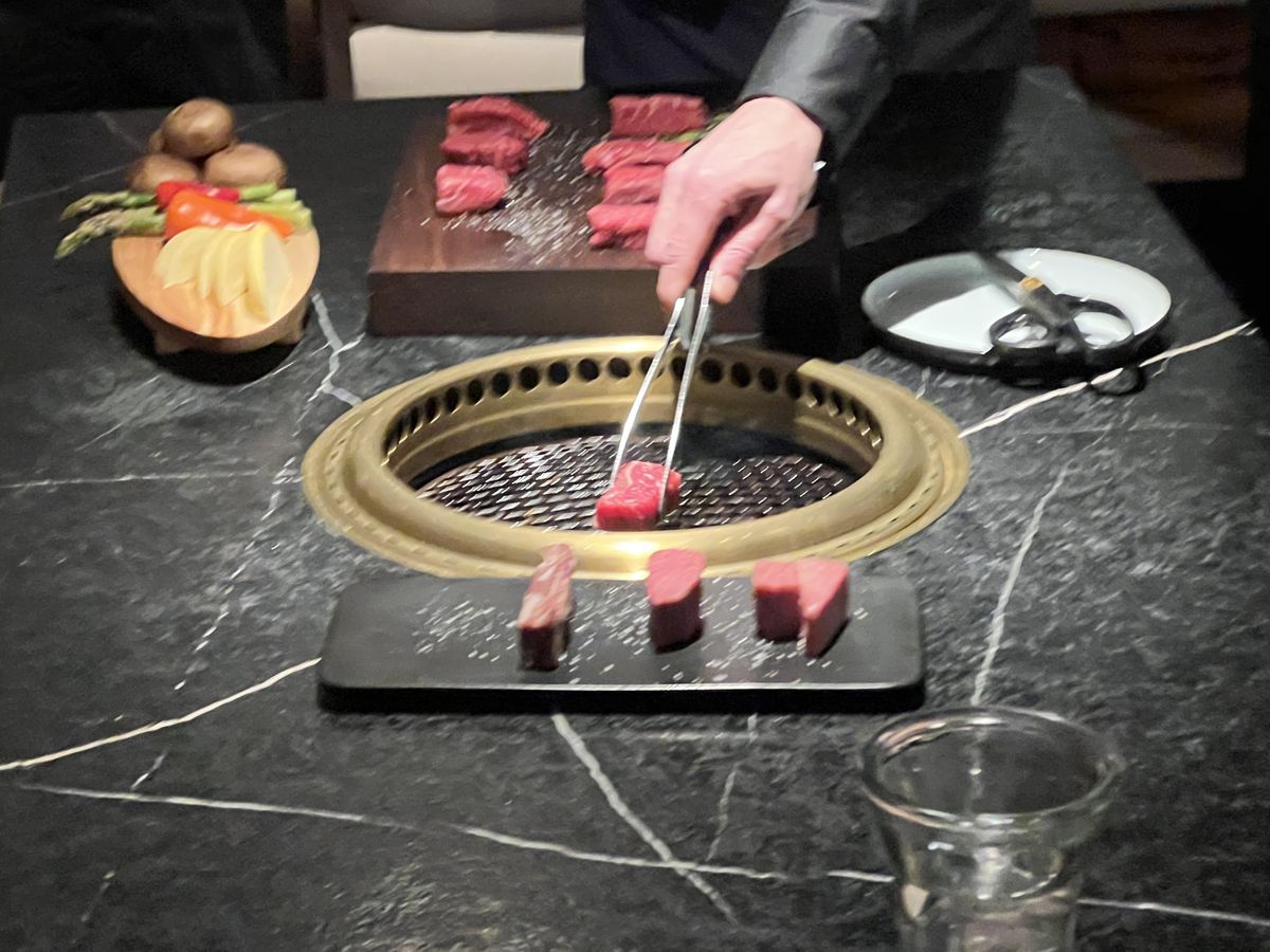 A hand holding tongs over a table grill, with a piece of raw beef.