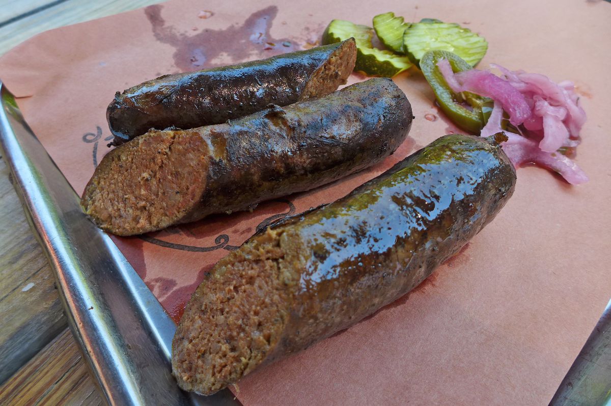 Three thick dark glistening links on brown paper with pickles and onions.