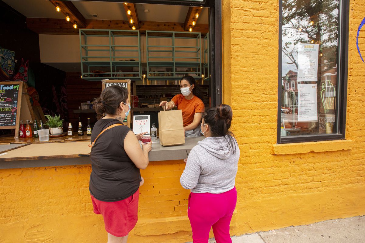 A server in a mask hands a large brown paper bag to two women wearing masks through a large walk-up window.
