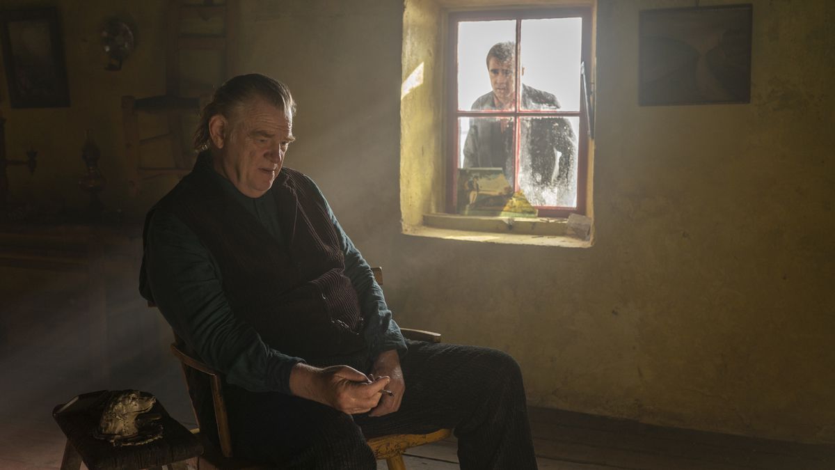 Brendan Gleeson sitting inside a house as Colin Farrell looks at him through a window in the film The Banshees of Inisherin