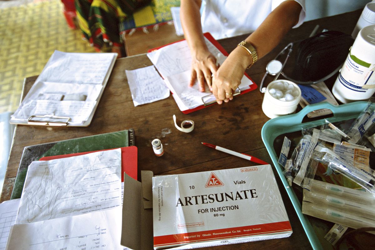 A medical worker prepares paperwork alongside documents and medical supplies including a box of injectable artesunate vials.