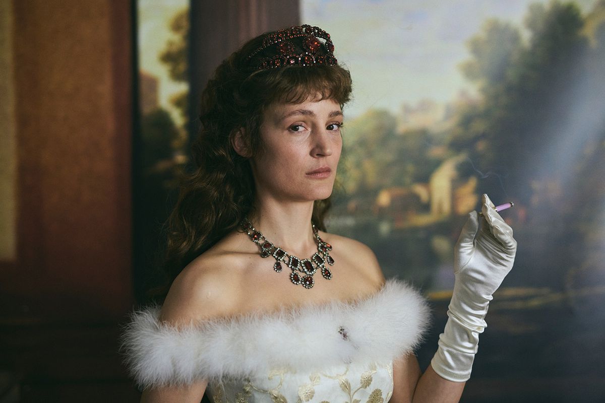 Vicky Krieps smokes a cigarette and wears a crown, a fancy dress, and a white glove in front of an ornate painting as Empress Elisabeth of Austria in Corsage.