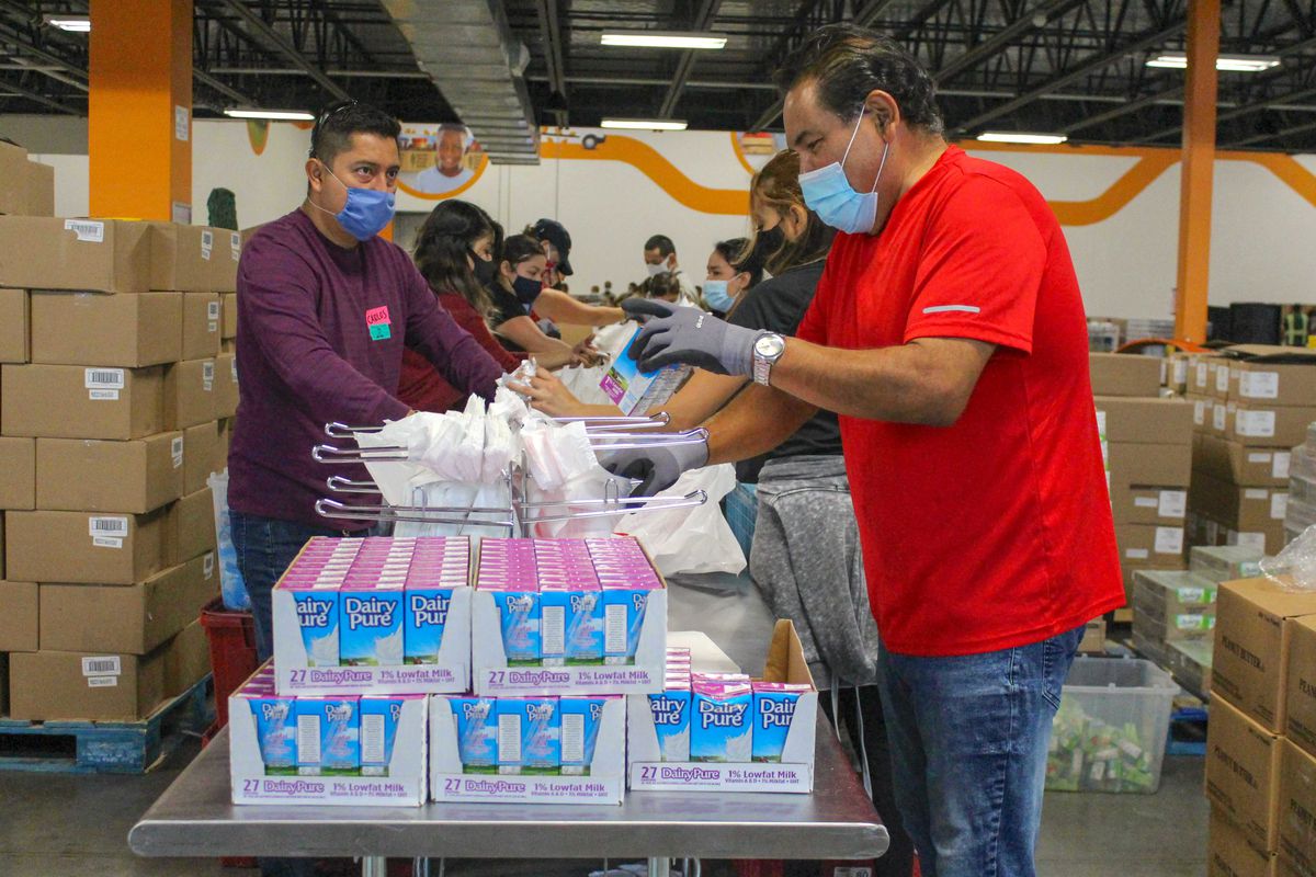 A man in a red t-shirt and blue face masks stands over boxes of shelf-stable milk, preparing to sort them into bags. 