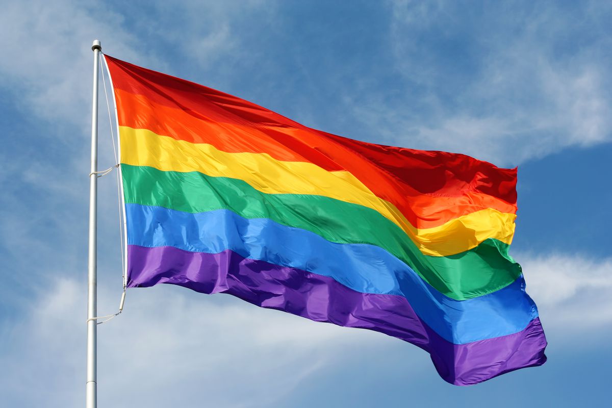 A rainbow flag blowing against a blue sky with faint white clouds. 