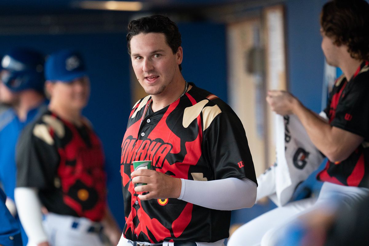 A man wearing a jersey that contains a giant face of Star Wars character Darth Maul stands in a baseball dugout holding a small water cup.
