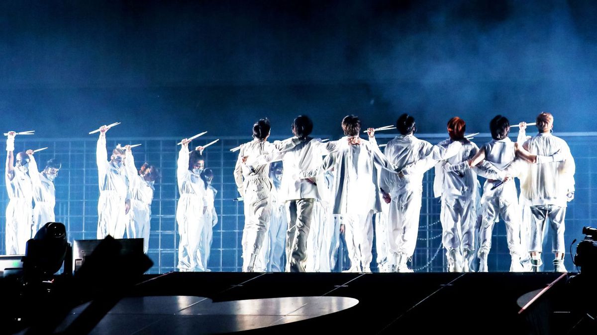 BTS members are dressed in white with their arms around each other looking from behind at their Permission to Dance on Stage concert in Los Angeles.  The dancers in the background stand with their hands in the air.  Everyone is holding drumsticks.