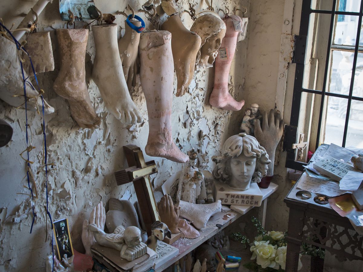 A wall with peeling white paint has multiple fake human limbs hanging from a rope on it. There is a ledge with various objects on it including a cross and several statue heads.