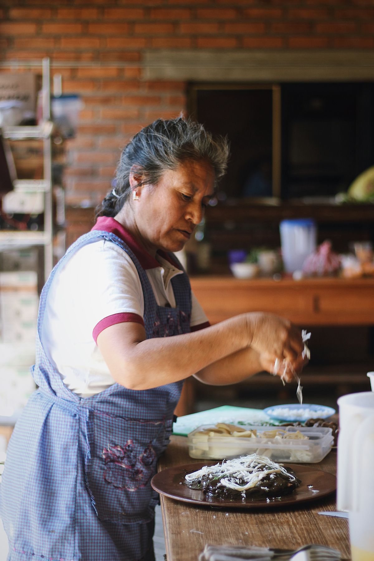 Woman in an apron sprinkles shredded cheese atop a dish containing mole negro.
