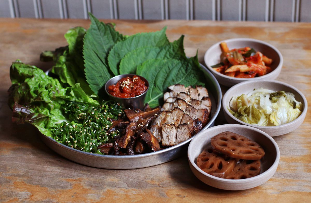 A round metal platter covered in sliced pork, perilla leaves, lettuce leaves, with some banchan on the side.