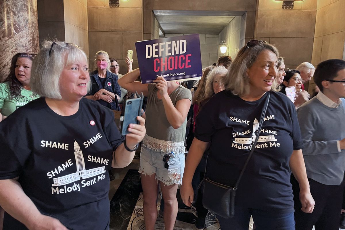 A crowd of people stands in a room. One person holds a protest sign that says “Defend Choice.” Two women, Pat and Neal, stand at the forefront, wearing matching black T-shirts printed with the words “Shame, Shame, Shame, Shame: Melody Sent Me”