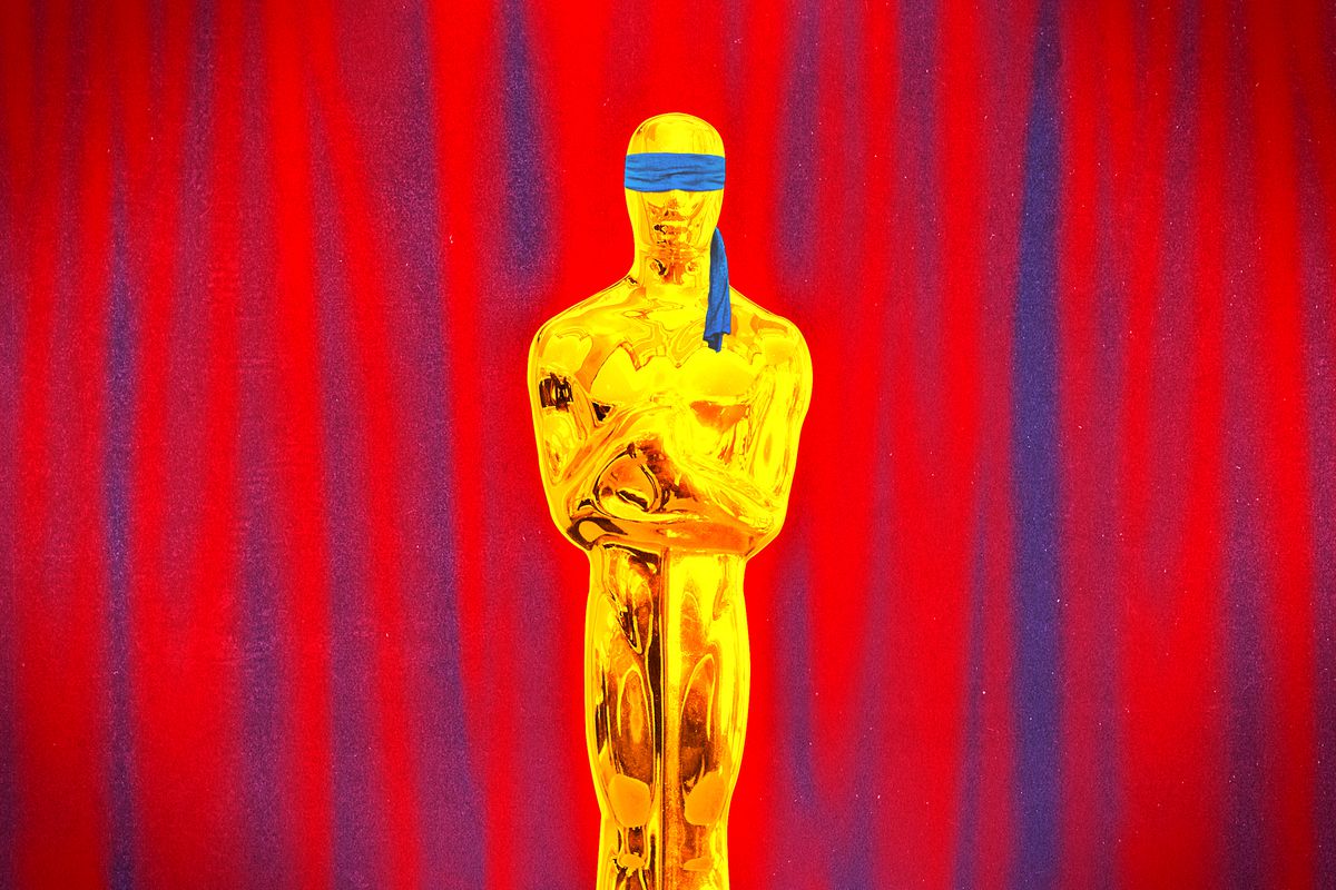 A blindfolded human-sized Oscars statue standing in front of a red curtain