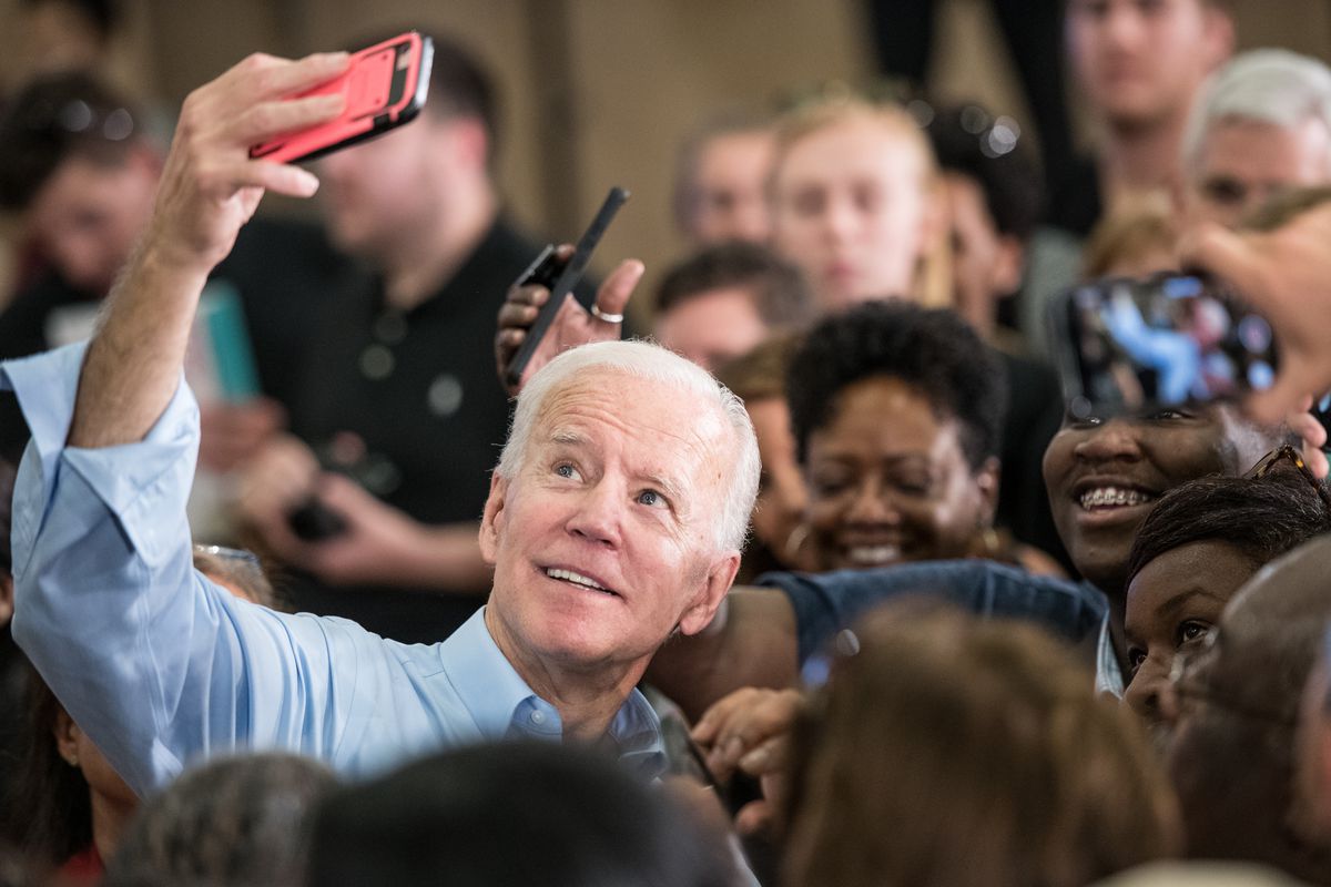 Biden raises a phone in a red case, smiling as supporters gather around him.