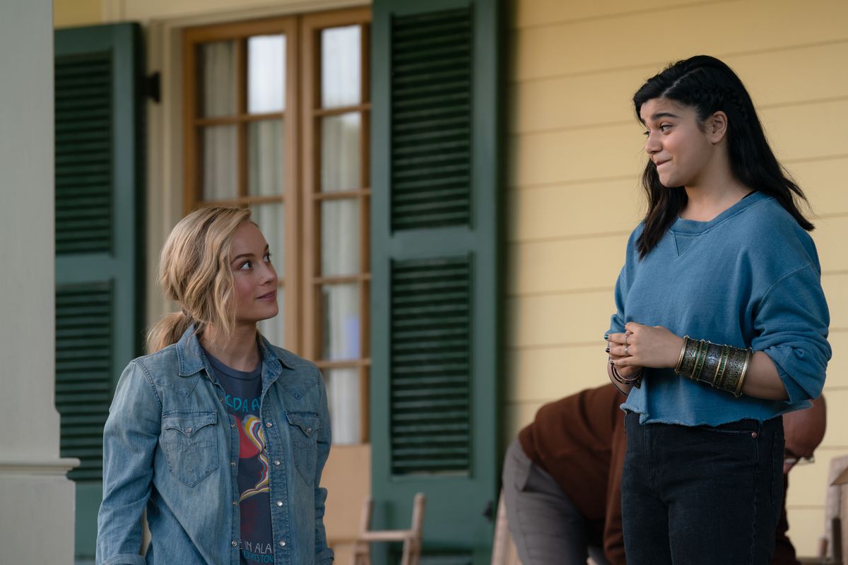 Brie Larson as Captain Marvel/Carol Danvers and Iman Vellani as Ms. Marvel/Kamala Khan stand outdoors on a porch and smile at each other in the Marvel Cinematic Universe movie The Marvels