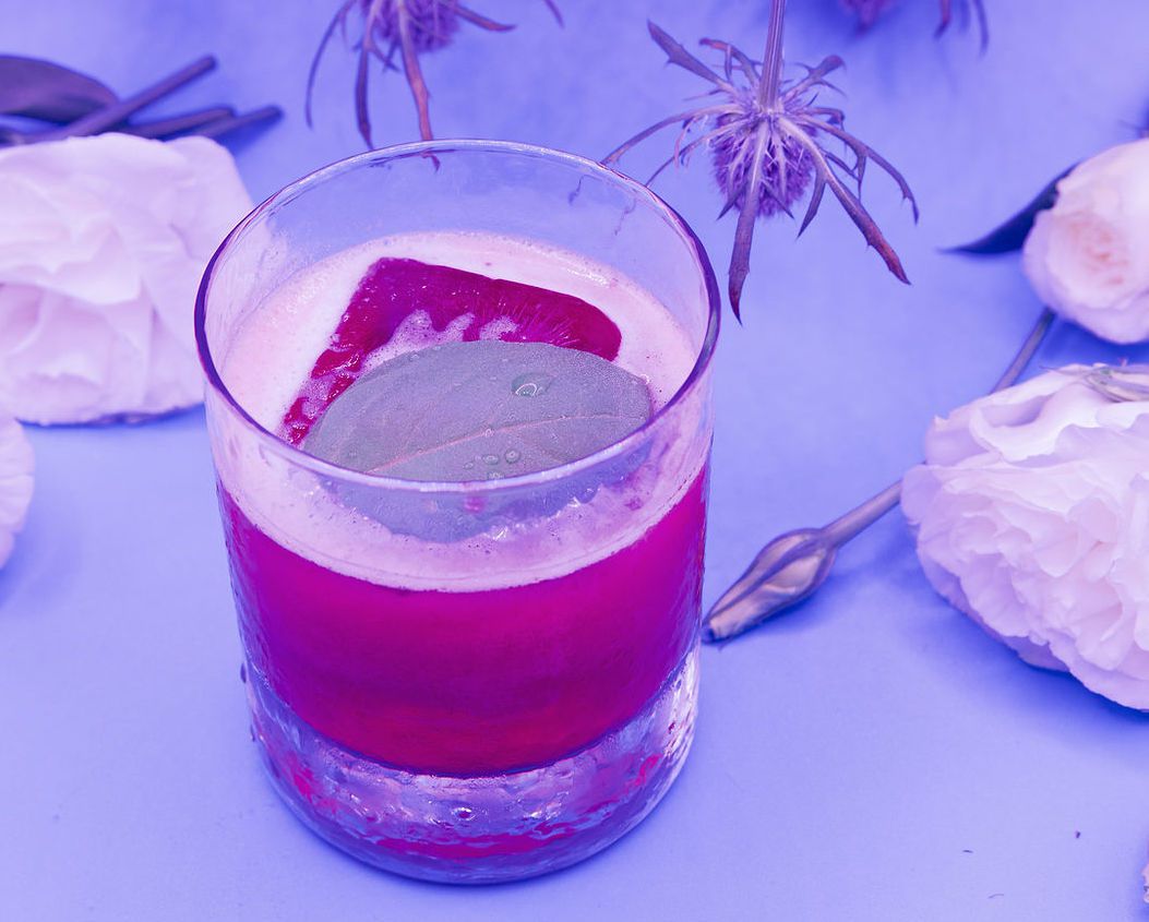 A red mocktail garnished with an ice cube and basil leaf against a purple background.
