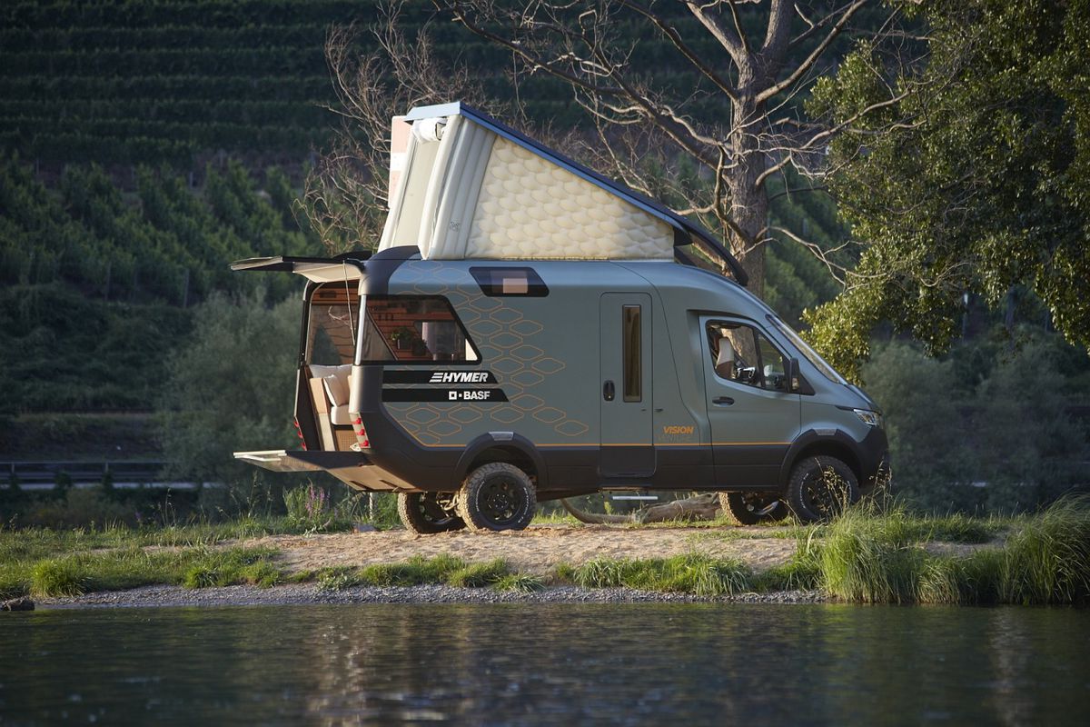 A modern camper van sits by a lake. The camper van boasts an open back door with small deck and a white pop-up sleeping area on the roof.