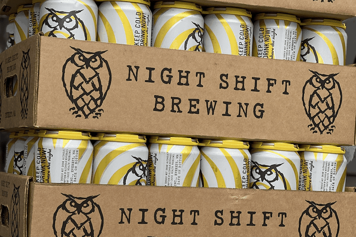 A stack of beer cans inside a cardboard container. The container reads, “Night Shift Brewing,” and the cans are adorned with a graphic owl logo.