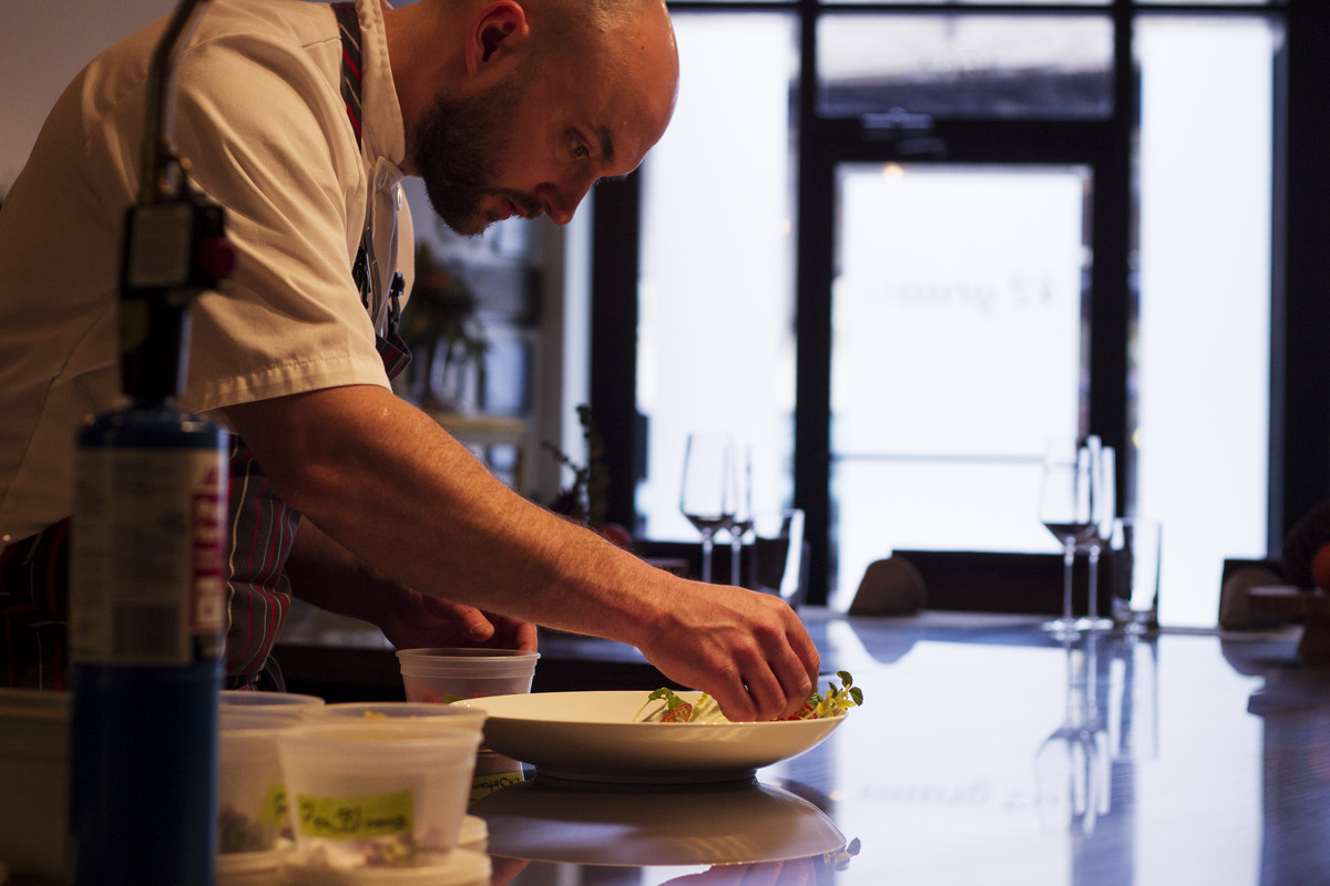 A chef bent over, plating food into a shallow bowl.