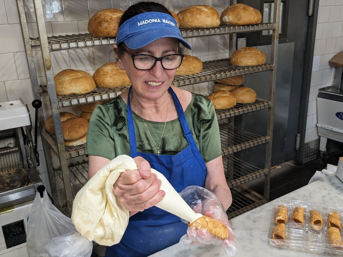 A woman in a blue apron fills a small pastry shell with a pastry bag.