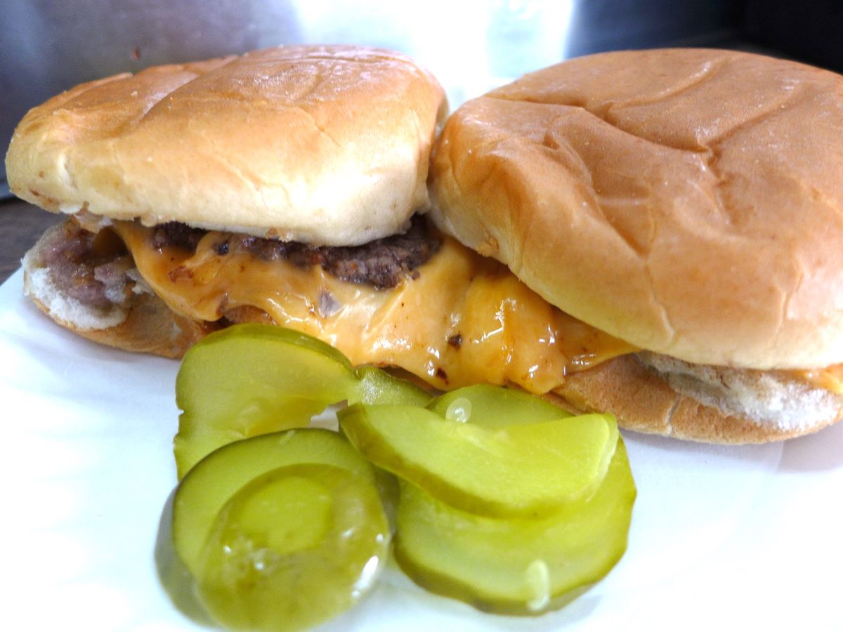 Two burgers strewn out on a paper plate, besides a side of neon green pickles.