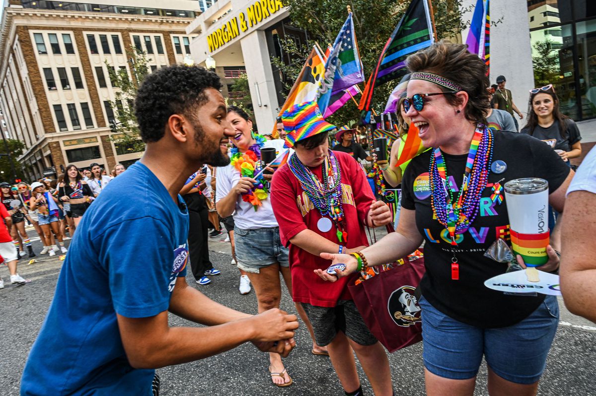 Frost, in a blue T-shirt, sporting a neat beard and a small afro, smiles as he hands a woman wearing colorful beads over a black Pride T-shirt and campaign button. She seems to be greeting him enthusiastically, Pride flags flying behind her.