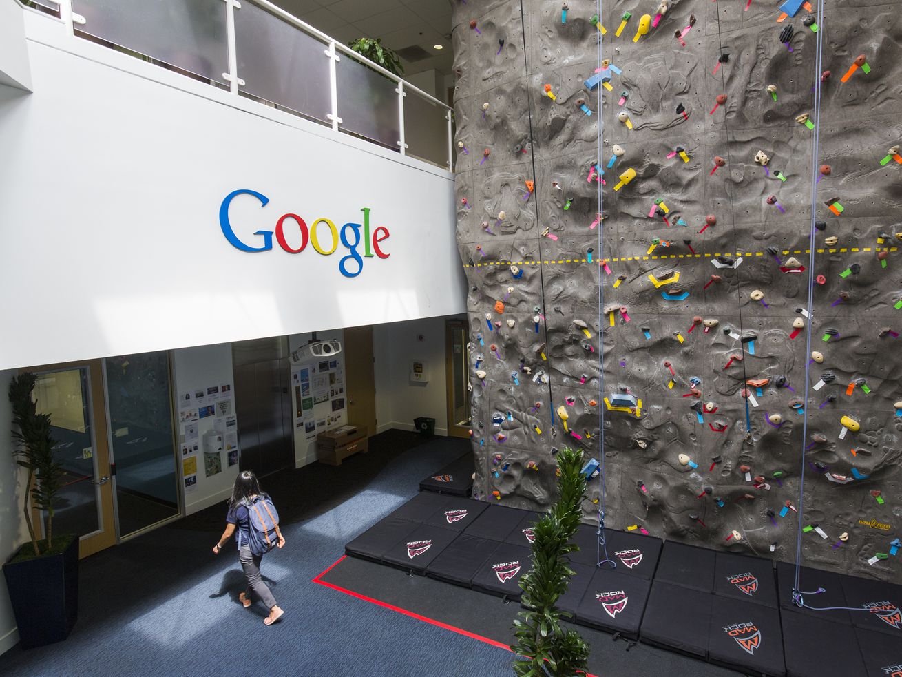 The inside of Google’s corporate headquarters, showing an indoor rock-climbing wall, with no one using it, and a person exiting.
