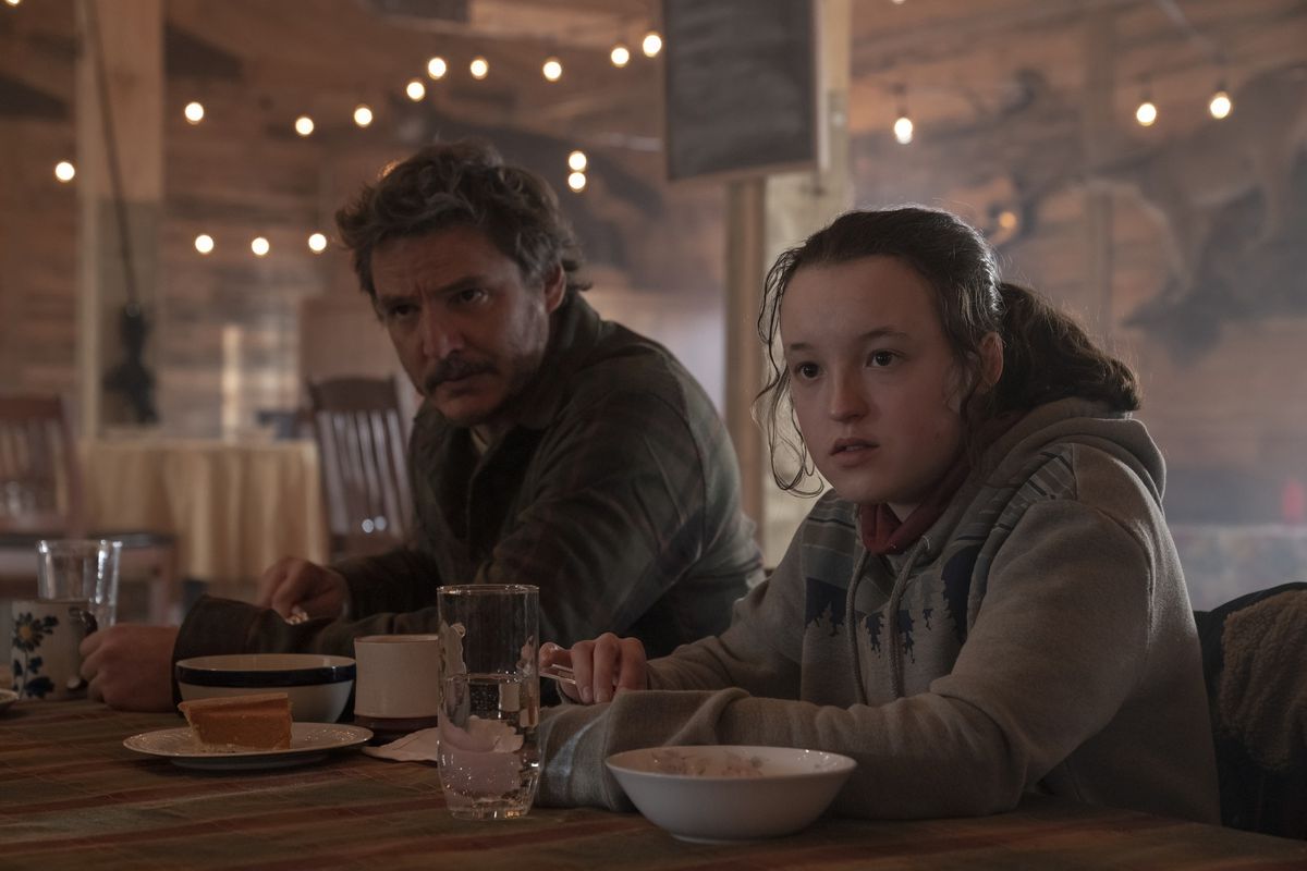 Pedro Pascal and Bella Ramsey sit together at a table with food in The Last of Us.