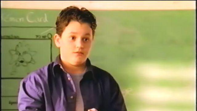 A still from a video showing Penn Badgley standing in front of a chalk board. He’s holding his hands together as he looks out towards the class. 